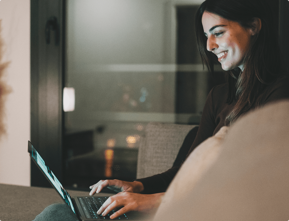 A brunette woman is sitting on a couch using her laptop. She has used her VPN to change her IP address, making her safer as she browses.