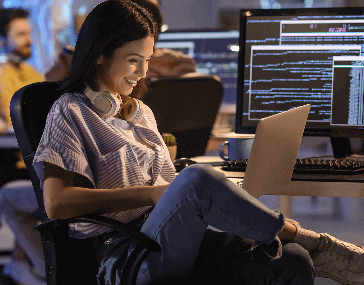 A woman wearing white headphones is sitting back in a chair. She is smiling at a laptop in her lap and has a desktop on her desk.