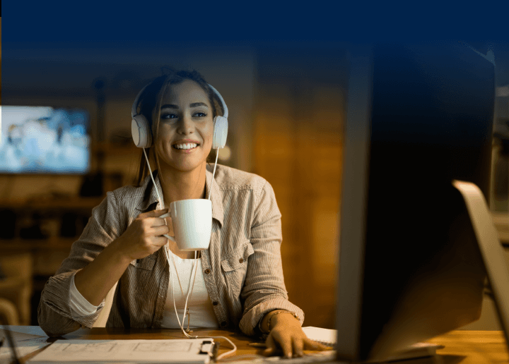 A woman sits at a computer wearing headphones and drinking from a mug. She is browsing Usenet with UsenetServer for the most secure and reliable Usenet access.